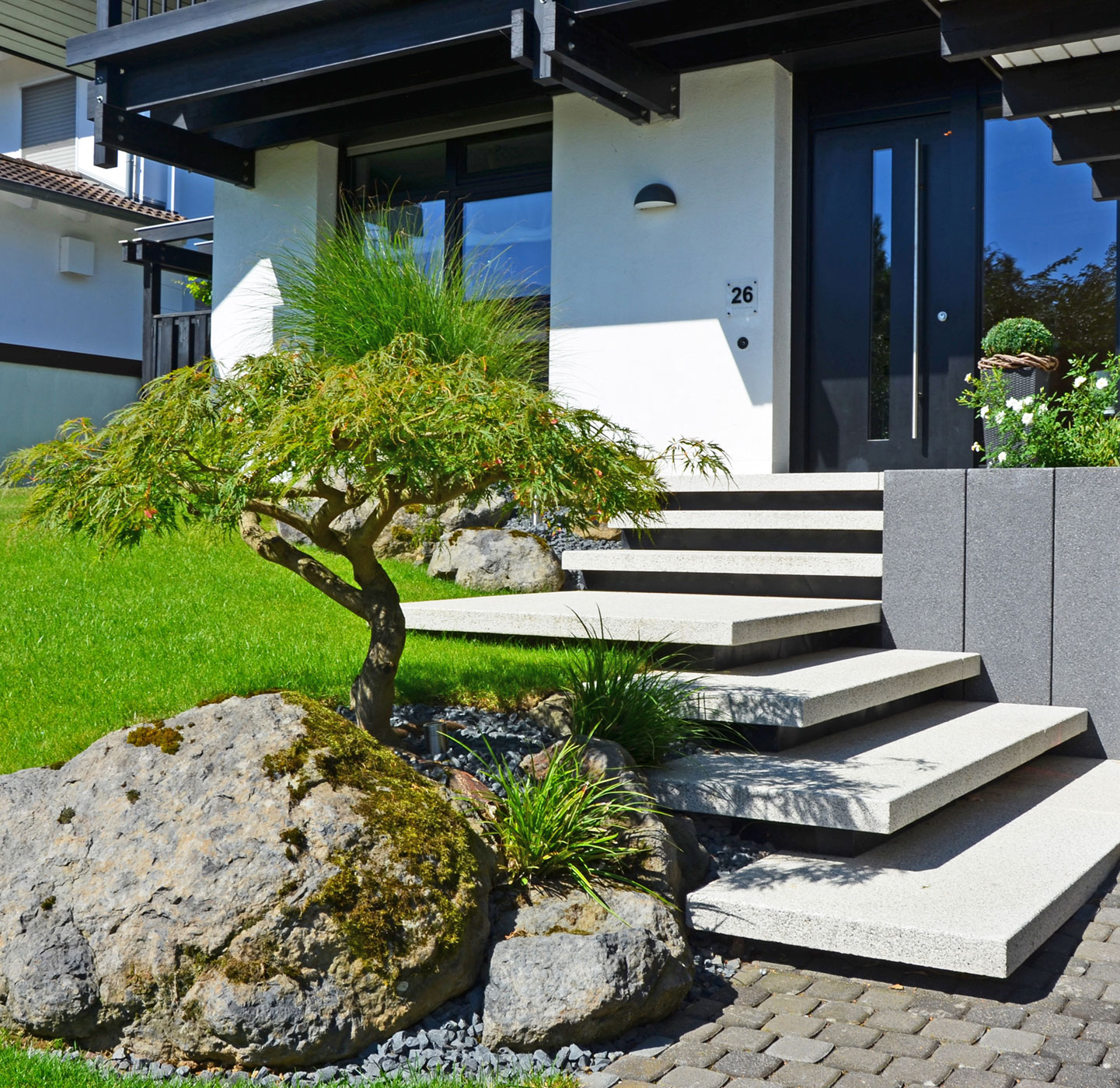 Treppe im Eingangsbereich eines Hauses mit einem schönen Vorgarten bestehend aus Steinen, einem kleinen Baum und Rasenfläche.