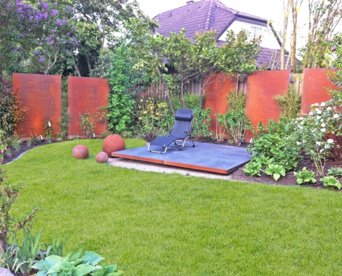 Garten mit einer grünen Wiese, ein Plattform mit einem Liegestuhl und einem Sichtschutz aus rostigen Cortenstahlwänden.