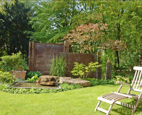 Sichtschutzelemente aus Holz und Metall sowie ein Wasserbecken in einem großen Garten , in dem ein schöner Holzliegestuhl steht.