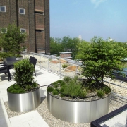 Moderne sehr große Dachterrasse einer Firma mit Sitzgelegenheiten und großen Chromkübeln, in denen Bäume und Büsche gepföanzt sind.
