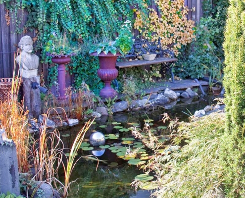 Wassergarten mit Steinbüste, klassischen Blumenkübeln aus Metall und einer Sitzbank.