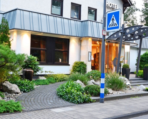 Moderner Hoteleingang mit gepflegtem Vorgarten durch immergrüne Bepflanzung mit dekorativen Gräsern und Zierkiefer.