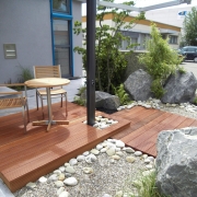 Holzterrasse mit Sitzmöbeln, Bekiesung und großen Steinen vor einem Haus.