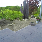Feinsteinzeugterrasse mit Blick auf eine Kiesfläche, Natursteinsäulen und grünen Pflanzen.