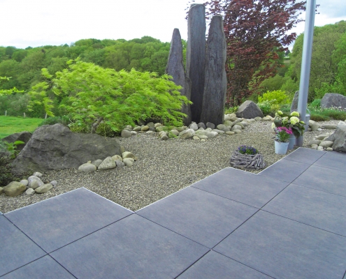 Feinsteinzeugterrasse mit Blick auf eine Kiesfläche, Natursteinsäulen und grünen Pflanzen.