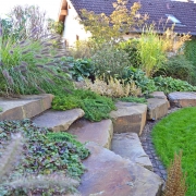 Aus Grauwacke erstellter Gartenhang mit einer Treppe auf eine andere Gartenebene und schöner Bepflanzung.