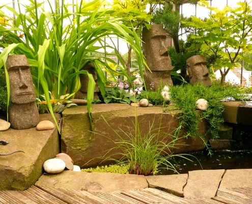 Wasserbecken mit grünen Pflanzen und Osterinsel Figuren auf einem Balkon.