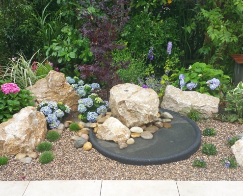 Steinbrunnen und Vogeltränke, blühenden Pflanzen, Kiesfläche und eine Sitzbank zur erholung im Garten.