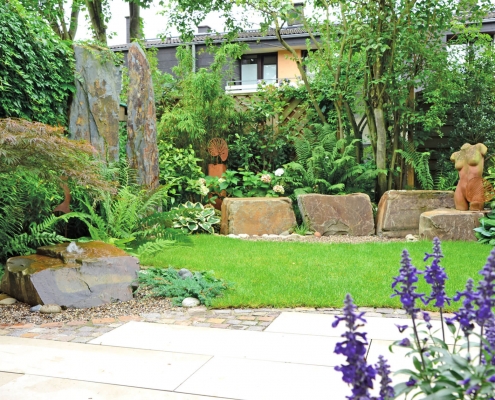 Garten mit Terrasse, Rasen, großen Steinstelen und einem Frauentorso aus Metall.